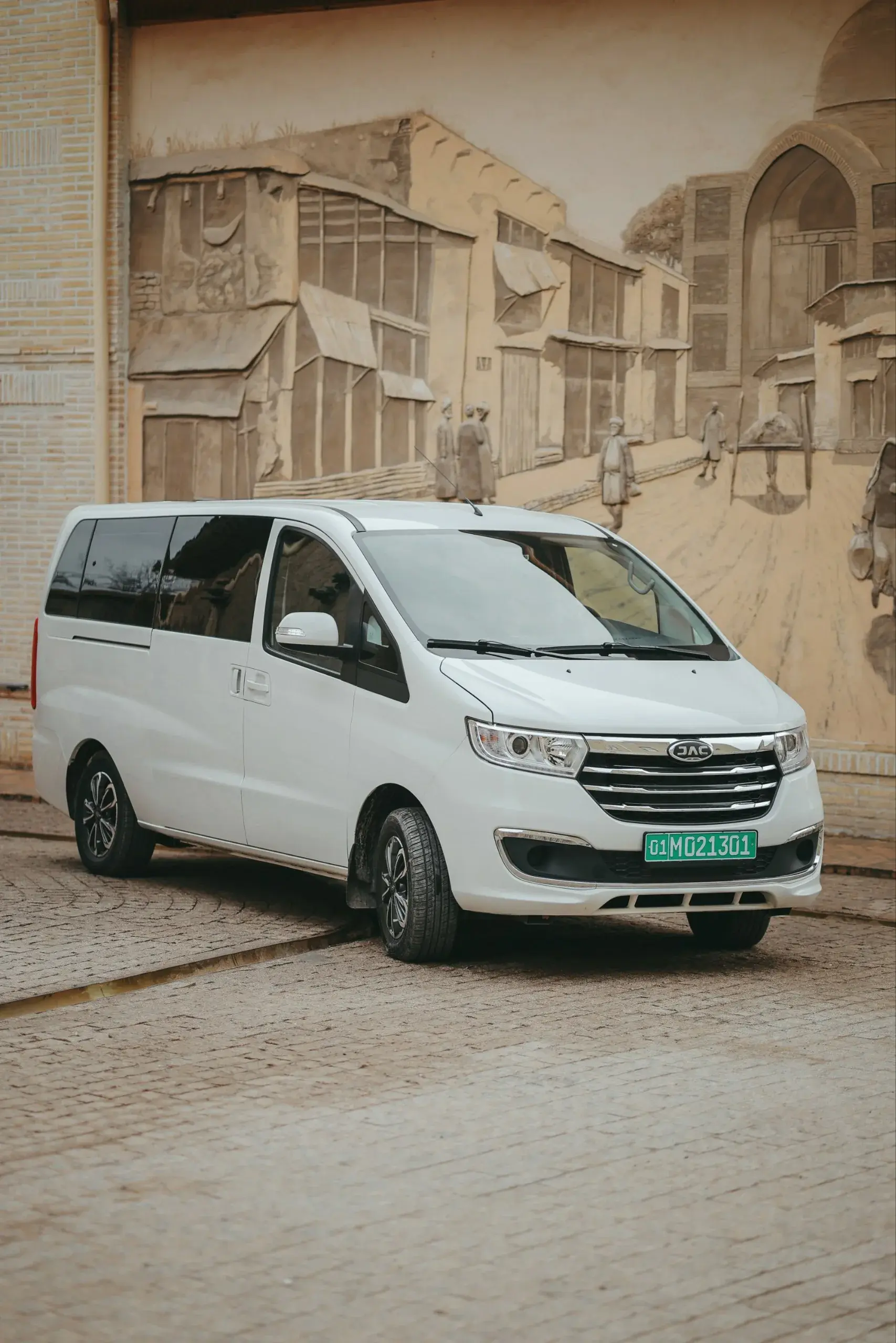 White JAC M3 Plus minivan in front of a carved old-city relief wall